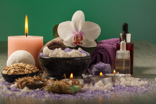 Spa composition with orchid flower, bowl with sea salt, bottles with aromatic oil, soap, scrub, candles and towels on wooden board and green background