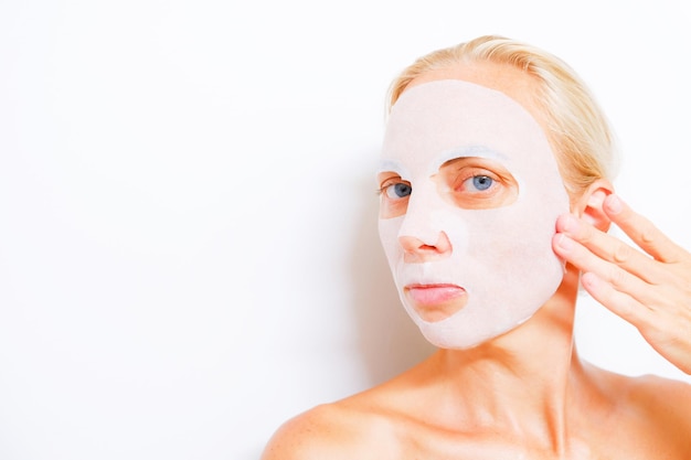 Spa girl with pleased facial expression applies mask on face gets beauty treatmentsstands with bare shoulders