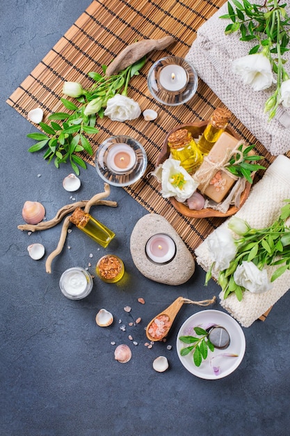 Spa still life wellness setting concept Assortment of essential oil cream sea salt natural soap candles white spring flowers and towel on a black table Top view flat lay