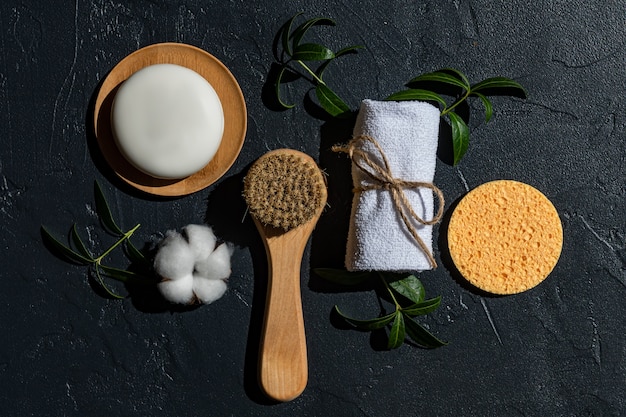 Spa treatment assessories on black background. Face massaging brush, cotton towel, natural sponge and handmade organic soap bar with herbal extract