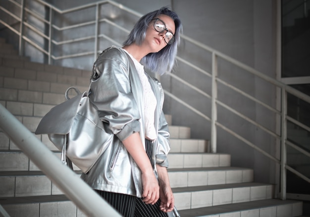 Space style girl with ashen blue hair is dressed in a cloak with a silvery fabric and a backpack on her shoulders posing on the street wearing glasses in the background of the steps of giving.