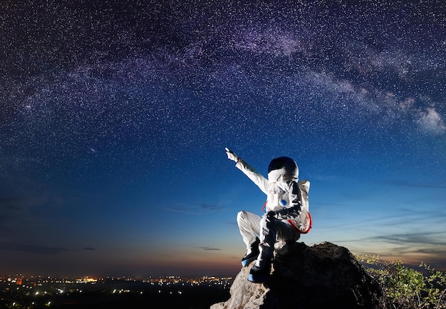 Space traveler sitting on rocky hill under fantastic starry sky