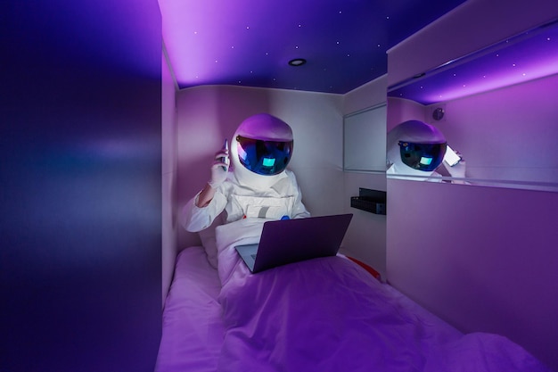 Spaceman work on a laptop and talking to a smartphone in capsule hotel sleeping module