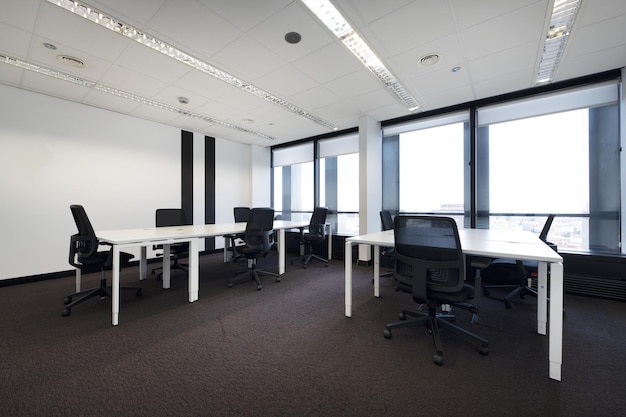 Spacious empty office with brown carpet and long white wooden tables with swivel black seats and technical ceiling and large windows with views