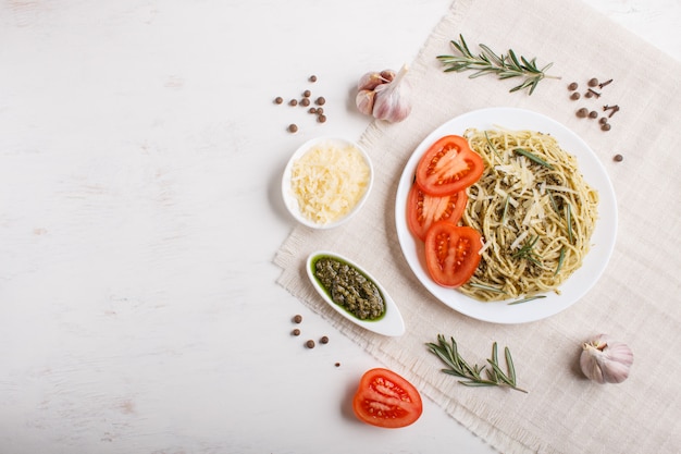 Spaghetti pasta with pesto sauce, tomatoes and cheese on a linen tablecloth on white wooden background.