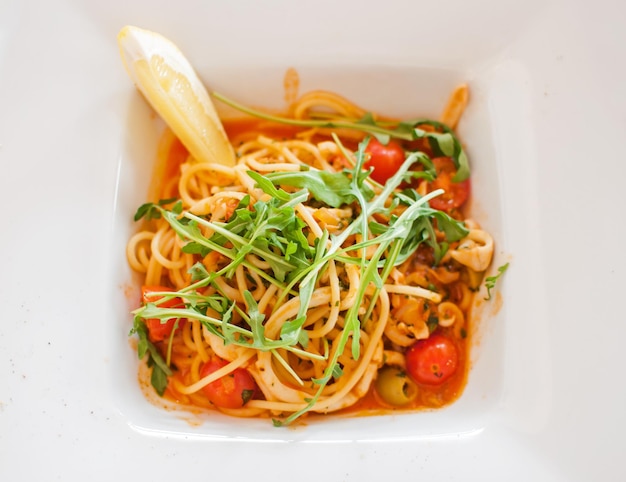 Spaghetti with squid and tomato and lemon arugula top view in white square plate