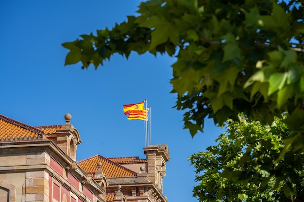 Spanish and Catalan flags on a pole waving on top of the historical building in Barcelona Spain Blue sky and green trees in a park