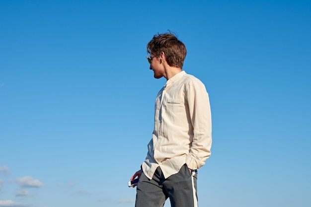 Spanish man in a beige shirt looking to the side with a phone in one hand, and the other in his pocket on clear sky