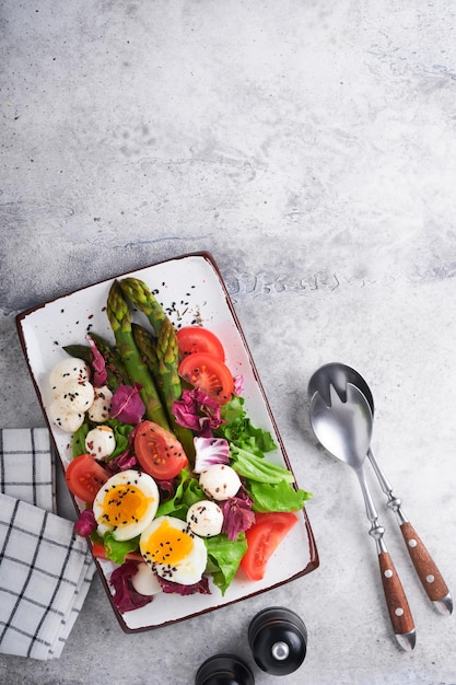 Sparagus tomato lettuce mozzarella black sesame flax oil olive salad and soft boiled egg on rectangular ceramic plate on grey concrete table background Healthy diet food concept Top view