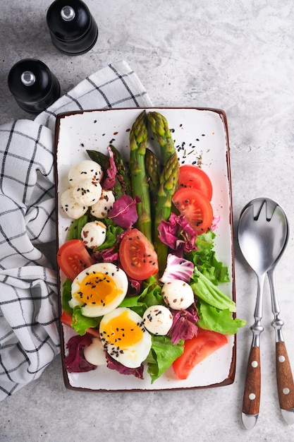 Sparagus tomato lettuce mozzarella black sesame flax oil olive salad and soft boiled egg on rectangular ceramic plate on grey concrete table background Healthy diet food concept Top view