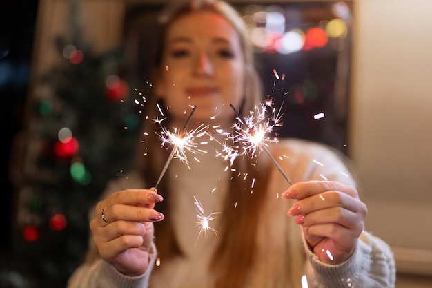 Sparklers bengal fire stick in female hands holding woman in knitted wool white cozy sweater on the background of a camping trailer house outdoor on new year and christmas eve night. sparkling sparks