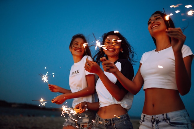 The sparklers in the hands of young girls on the beach. Three girls enjoying party on beach with sparklers. Summer holidays, vacation, relax and lifestyle concept.