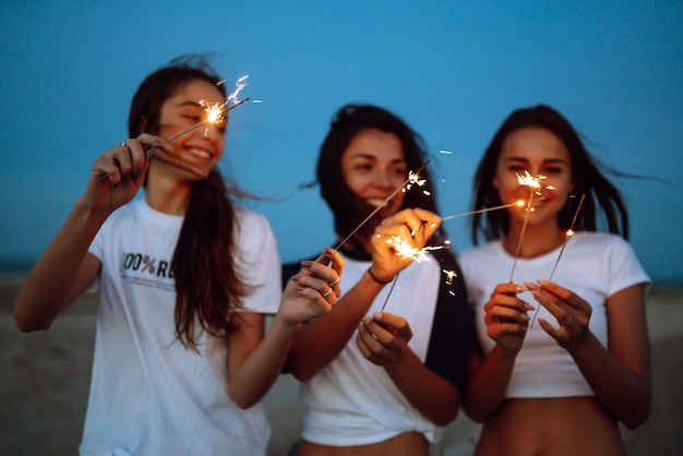 The sparklers in the hands of young girls on the beach. Three girls enjoying party on beach with sparklers. Summer holidays, vacation, relax and lifestyle concept.