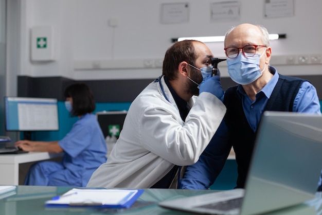 Specialist using otoscope to do ear examination on elder man, doing otolaryngology consultation with patient. Doctor with face mask examining infection to give prescription treatment.