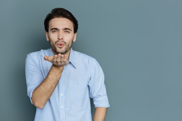 Specially for you. Pleasant nice young man sending you an air kiss and expressing his feelings while standing against grey background