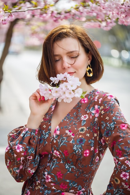 A spectacular woman in a bright dress stands against the background of sakura. A dark-haired woman in a beautiful outfit smiles on the street while walking