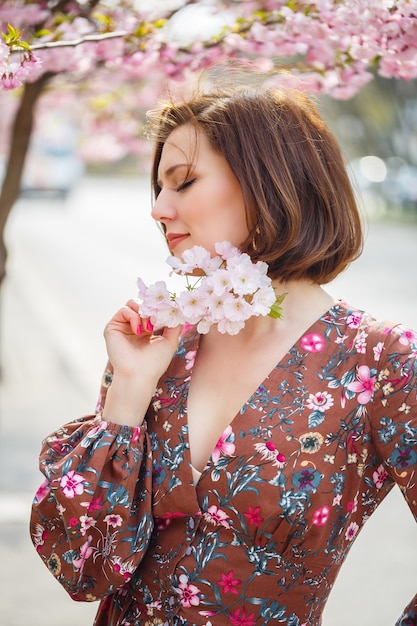 A spectacular woman in a bright dress stands against the background of sakura. A dark-haired woman in a beautiful outfit smiles on the street while walking