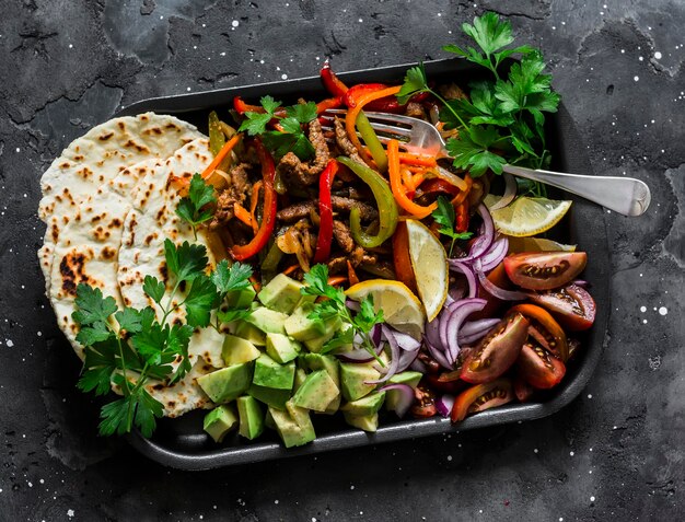 Spicy beef vegetables avocado corn tortillas fajitas on a sheet pan on a dark background