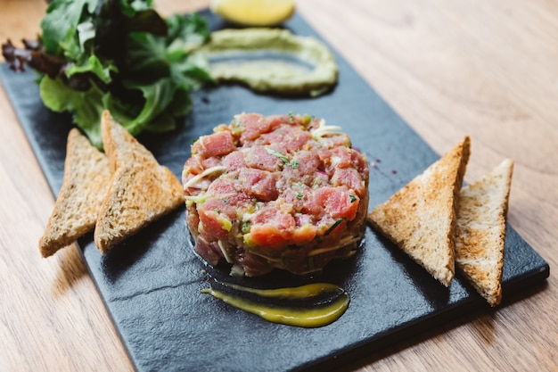 Photo spicy bluefin tuna tartare with sour and spicy sauce. served with toast and salad on black stone plate.