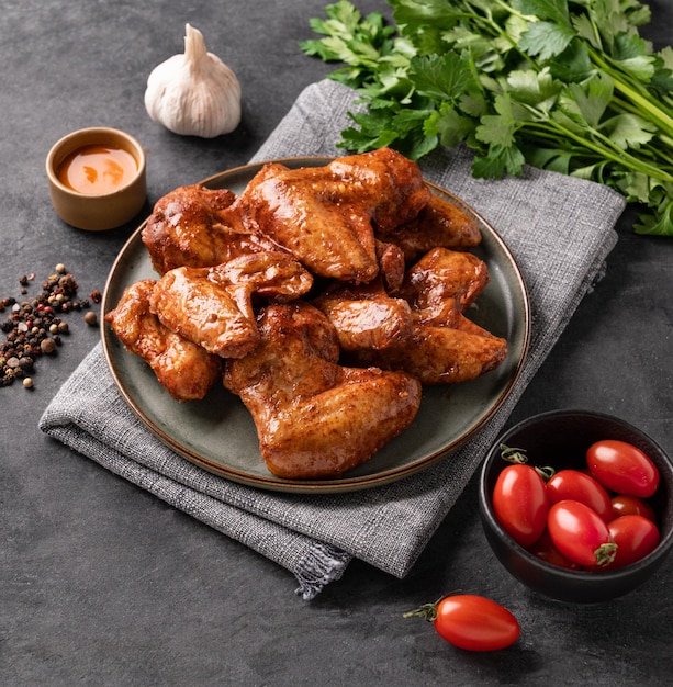 Spicy grilled chicken wings with sauce on a plate with tomato and herb on a dark background of slate or concrete