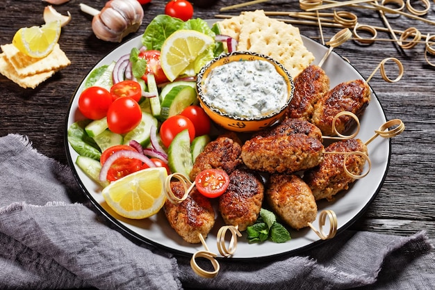 Spicy lamb and feta kofta skewers with fresh tomatoes, mint, red onion slices, crackers, and tzatziki sauce on a dark wooden table, top view, close-up