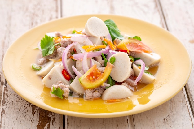 Spicy and sour fish ball salad with minced pork, tomato, carrot, red onion, shallot, celery, lime and chili in yellow ceramic plate on white old wood texture background, Thai food, Yum Look Chin Pla