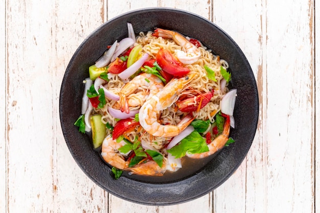 Spicy and sour instant noodle salad with shrimp minced pork tomato lime red onion shallot and celery in black ceramic plate on white old wood texture background top view Thai food Yum Mama