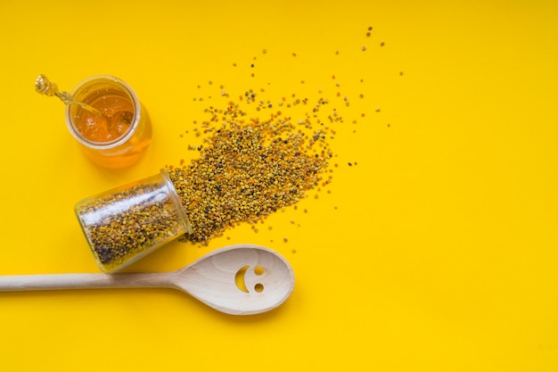 Spilled bee pollens; honey pot and smiley wooden spoon on yellow backdrop