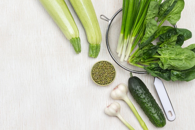 Spinach leaves and green onions in colander. Mash groats in green bowl. Zucchini and cucumber on table. White background. Flat lay. Copy space