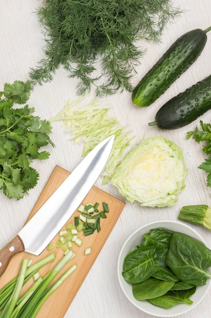 Spinach leaves in white bowl. Green onions and knife on cutting board. Cabbage, cucumbers and greens on table. White background. Top view