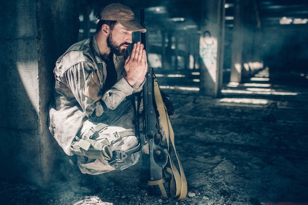 Photo spiritual man is sitting in squat position and praying. he is keeping his eyes closed and holding hands together close to face. also there is a rifle near his knees.