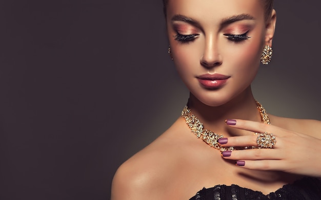 Splendid makeup with long black eyelashes and dark rose lipstick on the face of pretty woman french manicure on her nailsCloseup portrait of beautiful model dressed in a gilded jewellery set