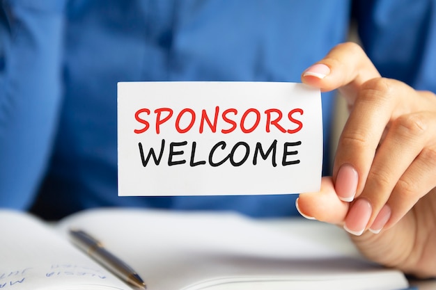 Sponsors welcome is written on a white business card in a woman's hand. Blue background. Business and advertising concept