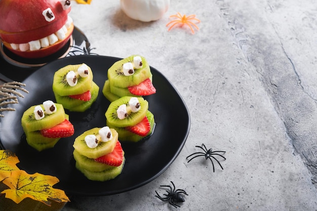 Spooky green kiwi monsters for Halloween Healthy Fruit Halloween Treats Halloween party kiwi strawberry apple and marshmallow monster on grey stone or concrete table background Selective focus