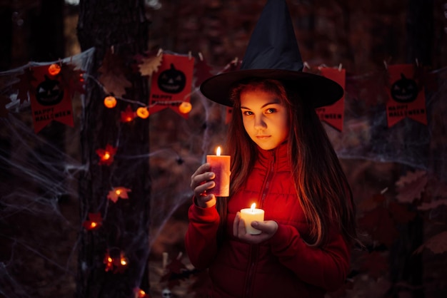 Spooky photo of small schoolgirl with red and white candle late in autumn forest Halloween celebration concept of witchcraft in forest