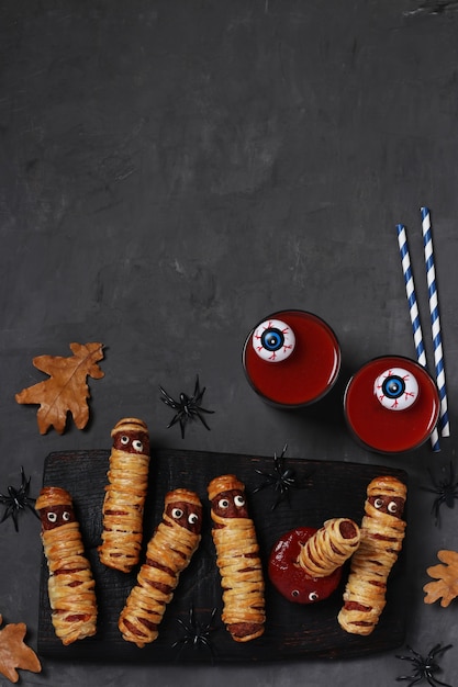 Spooky sausage mummies, tomato juice and sauce for Halloween party on dark wooden board. Top view. Flat lay. Vertical format. Copy space.