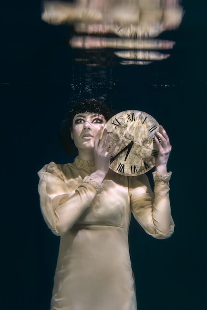 Spooky woman with clock in her hands in black veil underwater