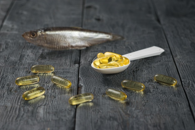 A spoon with fish oil capsules on the background of a small fish on a wooden table