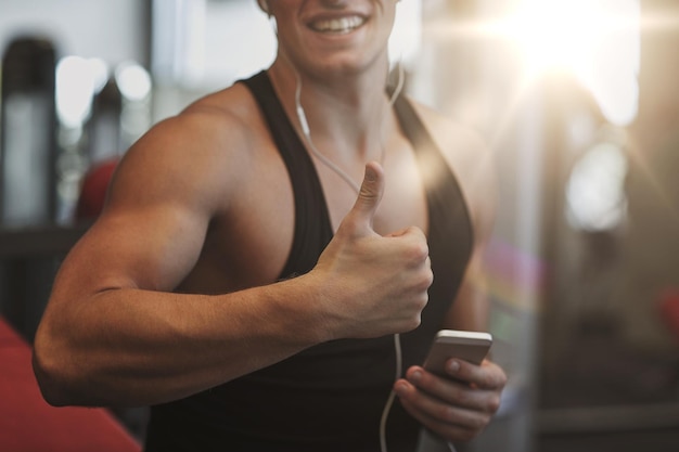 sport, bodybuilding, lifestyle, technology and people concept - happy man with smartphone and earphones listening to music and showing thumbs up in gym