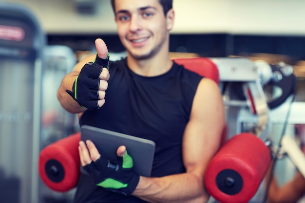 sport, bodybuilding, technology, gesture and people concept - happy young man with tablet pc computer showing thumbs up in gym