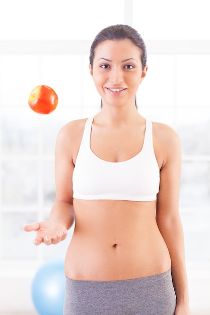 Sport and dieting. Beautiful young woman in sports clothing tossing an apple up and smiling at camera