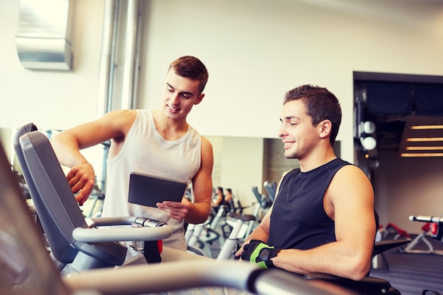 sport, fitness, equipment, technology and people concept - men with tablet pc computer exercising on gym machine