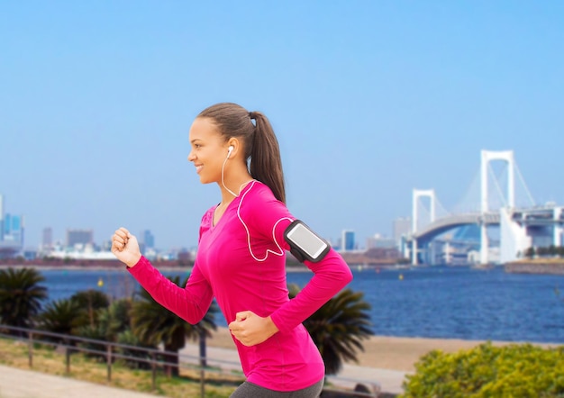 sport, fitness, health, technology and people concept - smiling young african american woman running with smartphone and earphones outdoors