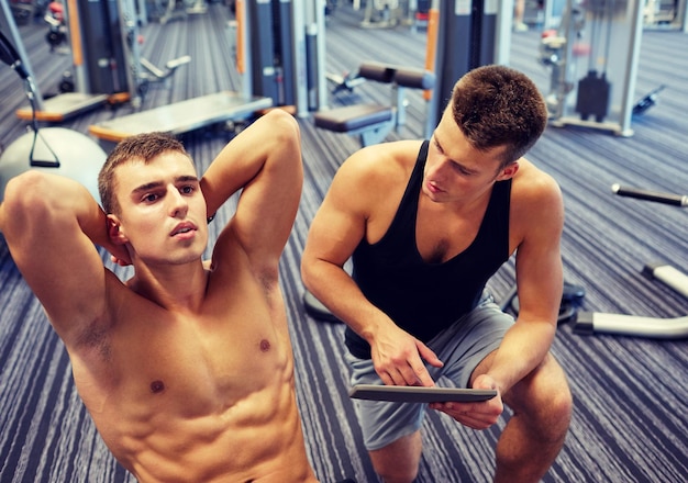 sport, fitness, lifestyle, technology and people concept - man and personal trainer with tablet pc computer and flexing abdominal muscles in gym