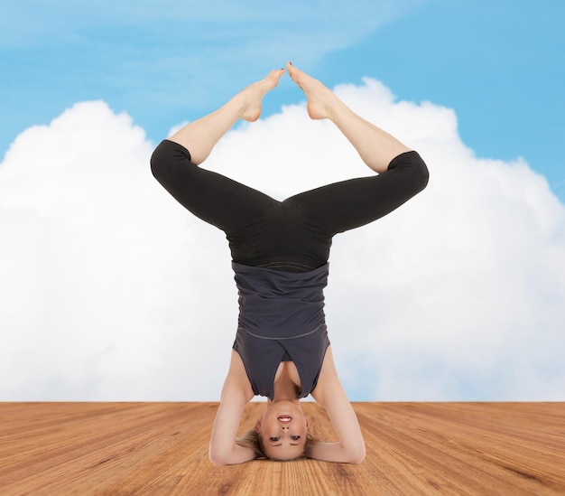 sport, fitness, yoga, people and health concept - happy young woman doing headstand exercise on wooden berth over white cloud and blue sky background