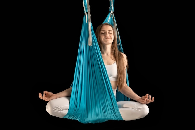 Sport girl practicing fly yoga in hammock and sitting in lotus position in the air Portrait of woman in hammock on black background