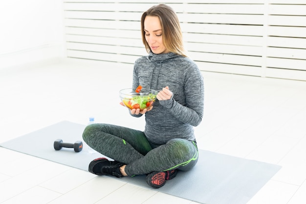 Sport, healthy lifestyle and people concept - young woman with salad and a dumbbell sitting on the