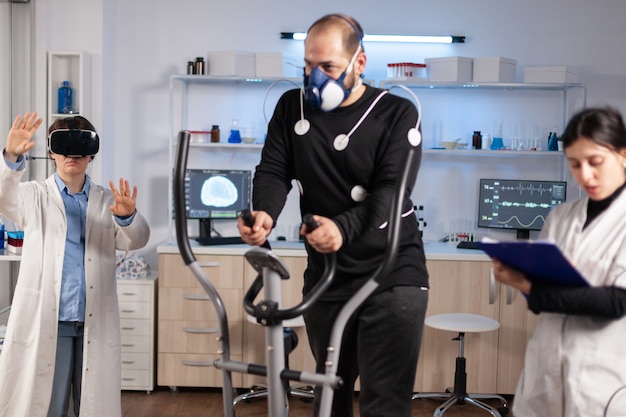 Sport performance scientist experiencing virtual reality goggles, athlete with electrodes running on cross training test. Measuring endurance with sensors.