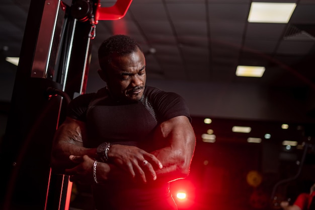 Sport portrait of african american athlete Strong bodybuilder man posing in gym and looking at his biceps Dark colors portrait