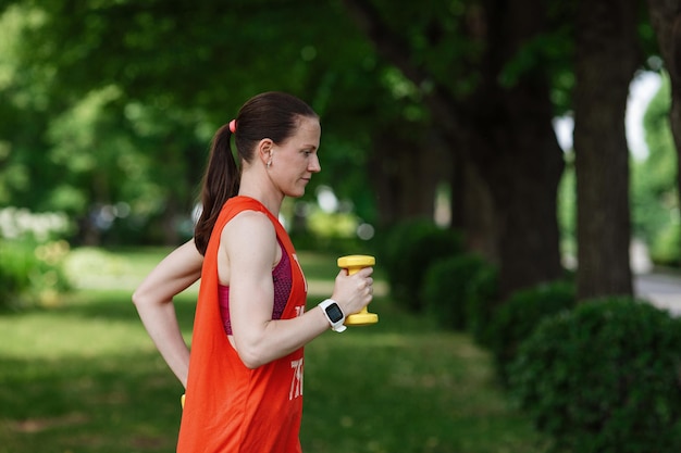 Sport training concept The girl is engaged in fitness in nature Side view of young woman doing stretching exercise using dumbbells Active athlete working out on fitness routine to lose weight